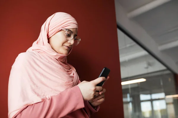 年轻的穆斯林女商人在办公室靠红墙时使用智能手机的侧视图肖像 复制空间 — 图库照片