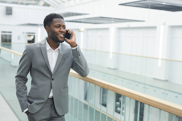 Waist up portrait of confident African-American businessman speaking by smartphone and smiling while leaning on balcony railing in modern office building interior, copy space