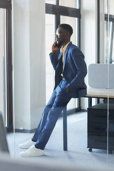 在现代办公室内靠桌子时 用智能手机讲话的成功非裔美国商人的全长侧面肖像 — 图库照片