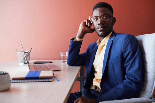 Portrait of young African-American businessman looking away while posing at workplace in office interior against red wall, copy space