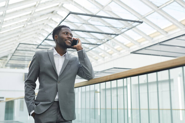 Low angle portrait of confident African-American businessman speaking by smartphone and smiling while leaning on balcony railing in modern office building interior, copy space