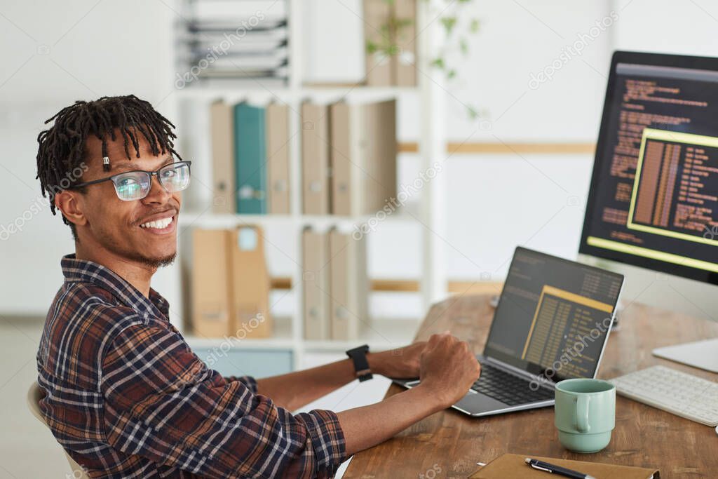 Portrait of smiling African-American IT developer looking at camera while typing on keyboard with black and orange programming code on computer screen and laptop, copy space