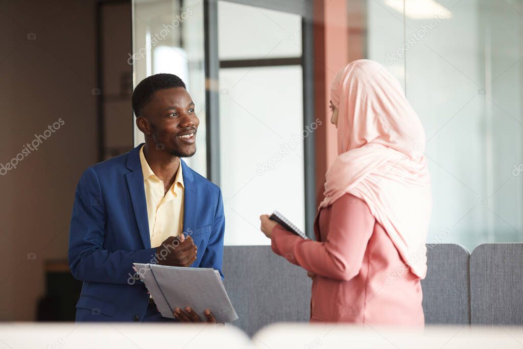 Side view portrait of smiling African-American man talking to young Muslim businesswoman and holding documents in modern office interior, copy space
