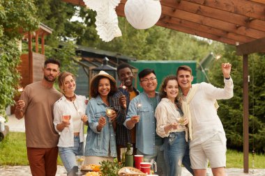 Çok ırklı bir grup genç elinde maytap tutarken ve kameraya gülümserken açık terasta yaz partisinin tadını çıkarırken,