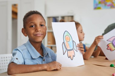 Uzay roketi resimlerini gösteren ve anaokulundaki veya çocuk gelişim merkezindeki resim derslerinin keyfini çıkarırken kameraya gülümseyen Afro-Amerikan çocuğun portresi.