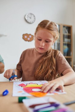 Okuldaki veya çocuk gelişim merkezindeki sanat ve zanaat derslerinden zevk alırken uzay roketinin resmini kesen kızıl saçlı kızın dikey portresi
