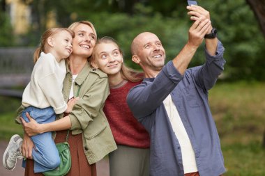 Modern mutlu ailenin belden aşağı portresi. İki kızı dışarıda fotoğraf çektirirken yeşil parkta yürümekten zevk alıyor.