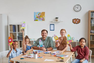 Uzay roketlerinin resimlerini gösteren çoklu etnik grupta gülümseyen erkek öğretmenin portresi, anaokulunda veya geliştirme merkezinde sanat ve zanaat dersleri alırken,