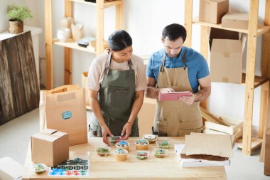 Yemek dağıtım servisindeki ahşap masada önlük takan iki işçinin yüksek açılı portresi.