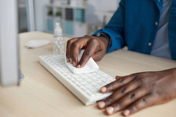 Close up of young African-American man wiping keyboard with sanitizing wipes while working at desk in post pandemic office, copy space