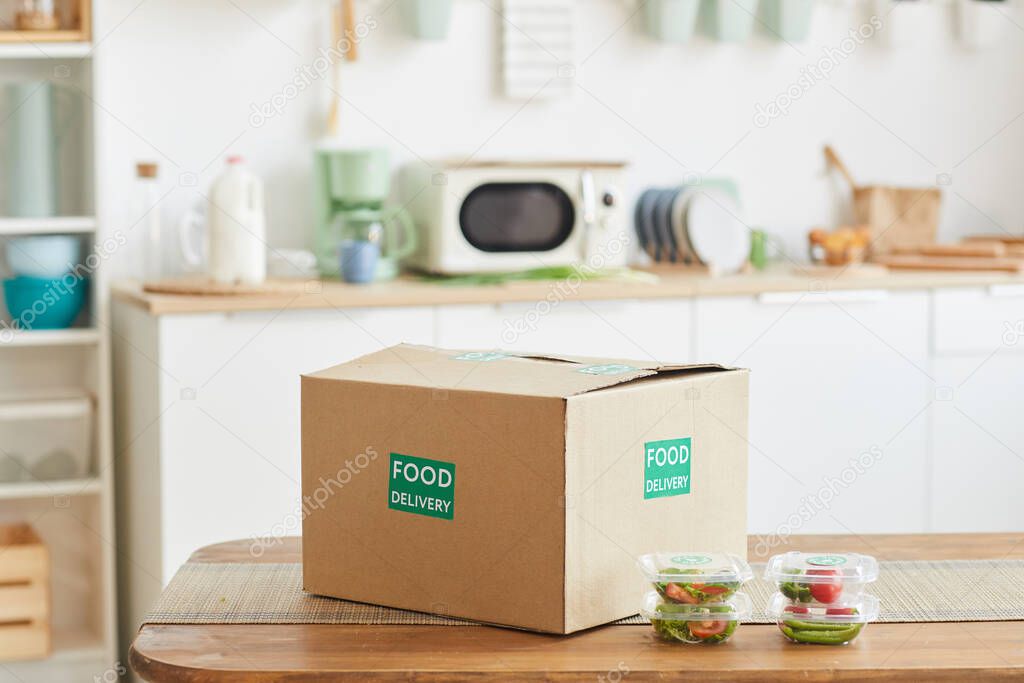 Background image of cardboard box with food standing on wooden table in white kitchen interior, food delivery service, copy space