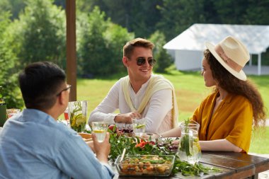 Gülümseyen genç çiftin renkli portresi. Yaz partisinde arkadaşlarıyla dışarıda yemek yerken birbirleriyle konuşuyorlar.