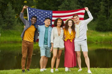 Amerikan bayrağı tutan ve yazın eğlenirken kameraya bakan çok ırklı arkadaş grubunun tam boy portresi.