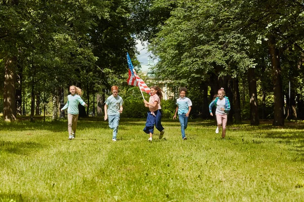 从广阔的视角看多民族孩子们奔向穿过绿色草坪的相机 女孩挥动着美国国旗 复制空间 — 图库照片