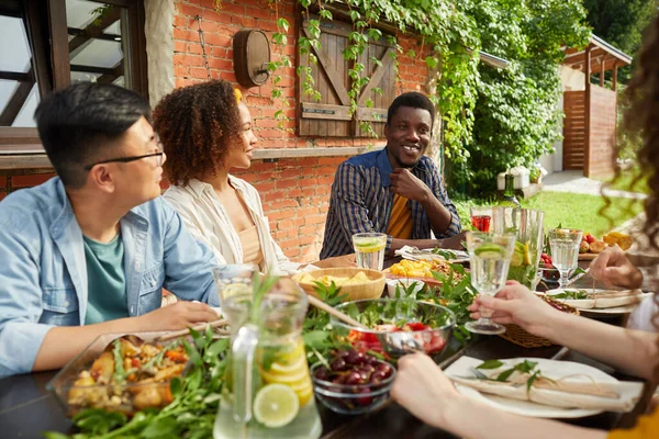 多民族朋友在露天平台上围坐在餐桌前享受晚餐的肖像 专注于微笑的非裔美国人分享故事 复制空间 — 图库照片