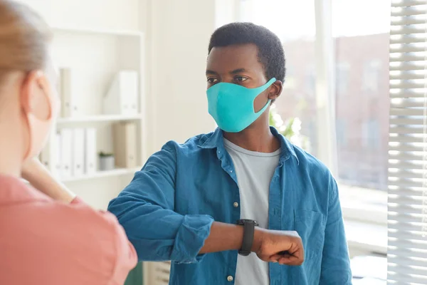 Waist up portrait of young African-American man wearing mask bumping elbows with colleague as contactless greeting while working in post pandemic office