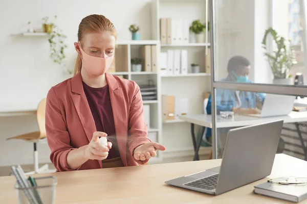 Portrait of young businesswoman wearing mask sanitizing hands while working at desk in cubicle at post pandemic office, copy space