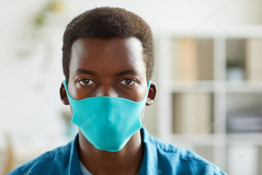 Head and shoulders portrait of young African-American man wearing mask and looking at camera while standing in post pandemic office, copy space