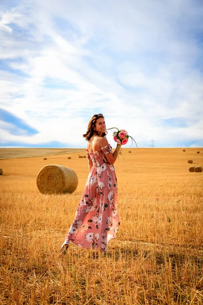 夏天礼服的年轻妇女站立与花束在收获的玉米地里与捆稻草 — 图库照片
