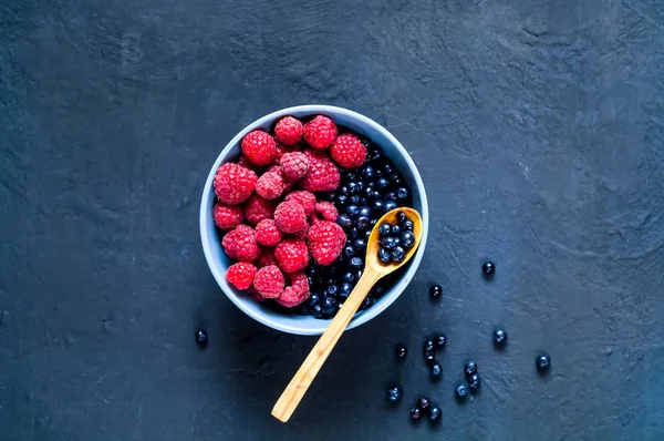 Houten lepel met blauwe bosbessen en frambozen in blauwe kom, het concept van biologische bessen op donkere achtergrond. Berry plukken, kopiëren van ruimte, close-up, banner — Stockfoto