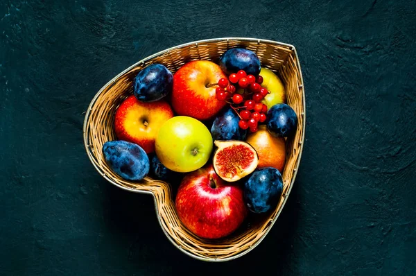 Red apples, plums and figs in straw vase made in the form of heart on dark blue background, top view, organic food concept, detox