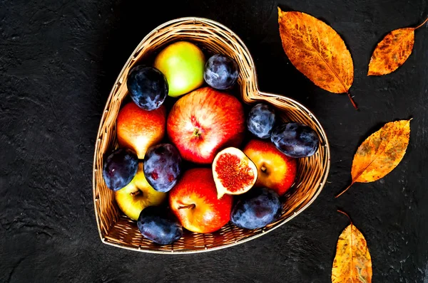 Red apples, plums and figs in straw vase made in the form of heart on dark blue background, organic food concept, clean food or vegetarian concept.Flat lay. Copy space.