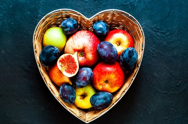 Red apples, plums and figs in straw vase made in the form of hea