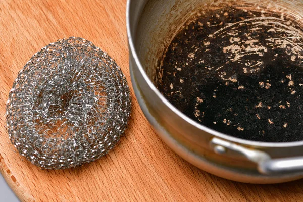 A spoiled burnt pan with a hard metal brush lies on a wooden Boa