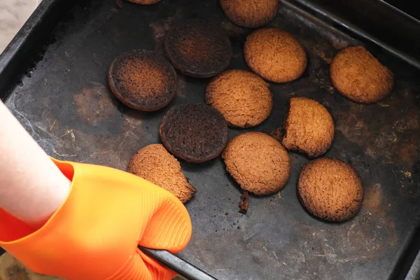 Burnt cookies. A hand in  orange oven glove, potholder picks up a black baking tray with burnt cookies