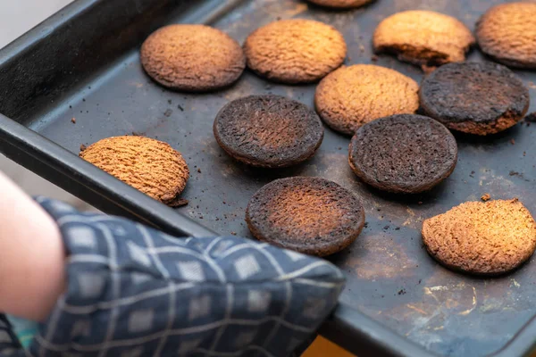 Burnt cookies. A hand in a oven glove, potholder picks up a black baking tray with burnt cookies