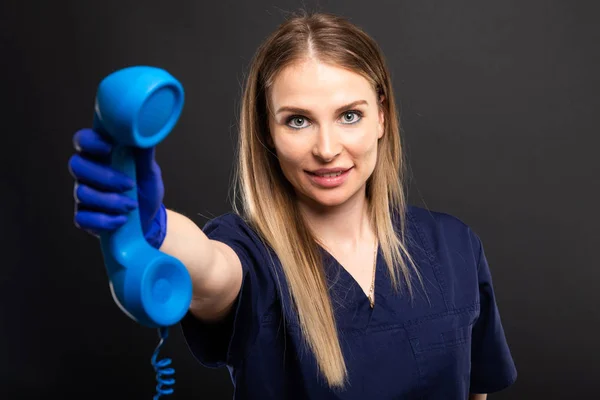 Female doctor wearing scrubs handing blue telephone receiver and smiling on black background