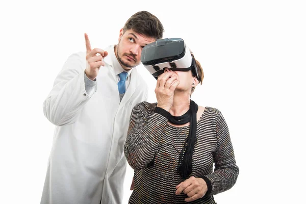 Male doctor pointing to female senior patient with vr goggles isolated on white background