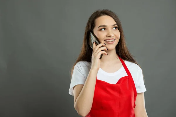 Portrait of young female supermarket employee talking at smartphone and smiling on gray background with copy space advertising area