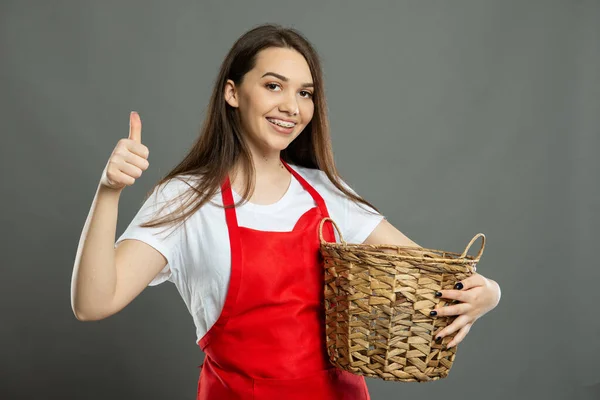Portrait of young female supermarket employee holding basket showing like on gray background with copy space advertising area