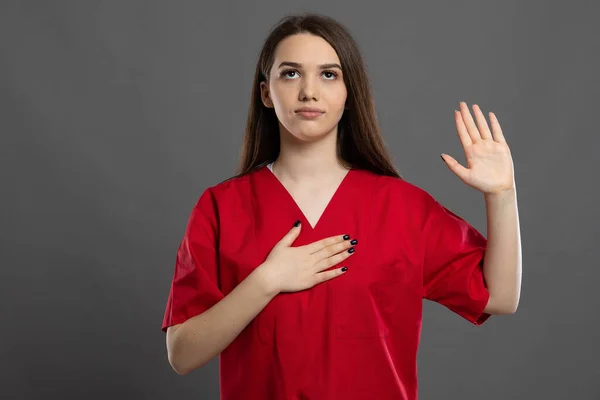Young graduate nurse pledging an oath wearing red scrubs on a grey background
