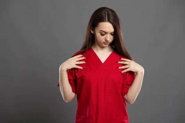 Long brown hair young graduate nurse checking her red hospital clothes on a grey background