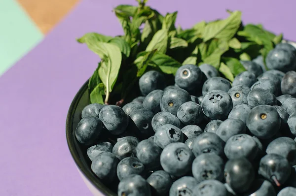 Fresh delicious blueberries with mint in bowl on purple background.