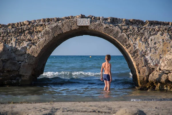 穿着泳装的年轻男孩站在一座古老的石桥下 在海边的水中 在希腊 — 图库照片