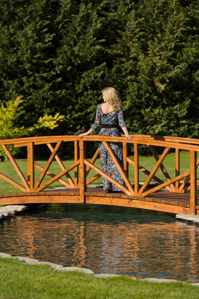 Beautiful long haired blonde woman stand on a wooden bridge over an artificial pond, in natural surround, green background.