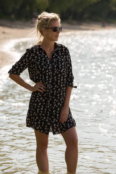 Blondýnka Stojí Vodě Pláži Krátkých Šatech Slunečních Brýlích Podsvícení — Stock fotografie
