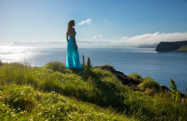 Oldukça uzun saçlı sarışın kadın bir kıyı tepenin üstünde uzun turkuaz elbise duruyor. İrlanda, doğal sahne, güzel manzara.