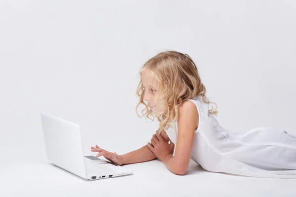 漂亮可爱的金发碧眼的小女孩 穿着白色的衣服 躺在明亮的工作室的地板上玩着白色的笔记本电脑 — 图库照片