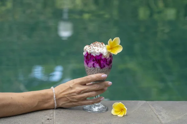 Dívka ruka drží sklenici s hia semena pudink na pozadí vody v bazénu. Chia semínka pudink s červený drak ovoce a bílého jogurtu ve sklenici k snídani — Stock fotografie