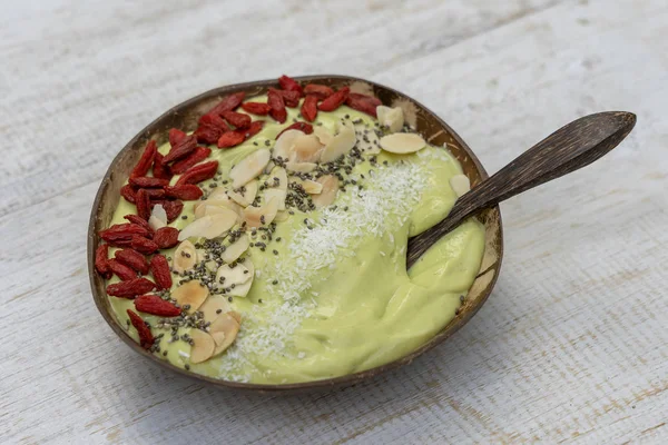 Smoothie verde em tigela de coco com abacate, bagas de goji vermelho, flocos de amêndoa, chips de coco e sementes de chia para o café da manhã, close up. O conceito de alimentação saudável, superalimento — Fotografia de Stock