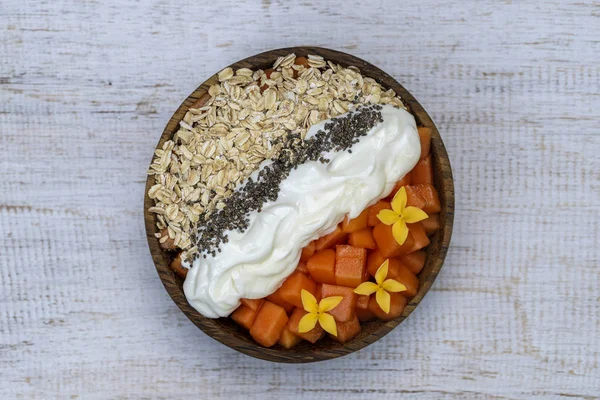 Rodajas de papaya dulce con copos de avena, semillas de chía y yogur blanco en un tazón de coco sobre fondo de madera blanca, de cerca — Foto de Stock