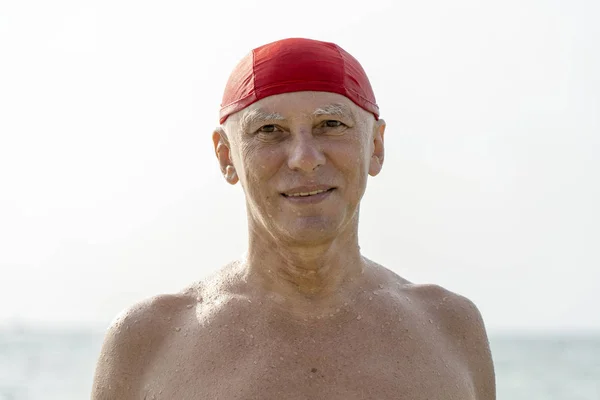 Oudere man in een rode zwemmen hoed op het strand in de buurt van het zeewater — Stockfoto