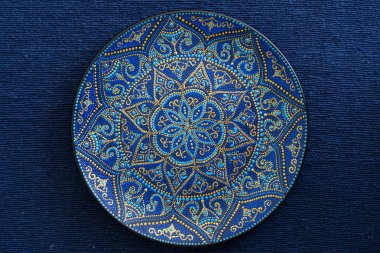Mavi ve altın renklerde dekoratif seramik plaka, mavi kumaş arka planda boyalı plaka, closeup. Akrilik boyalar ile boyanmış dekoratif porselen plaka, el işi
