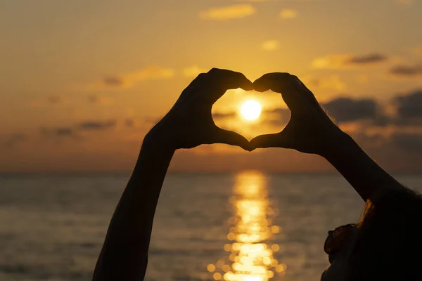 Mãos formando uma forma de coração com silhueta do pôr do sol perto da água do mar, de perto — Fotografia de Stock