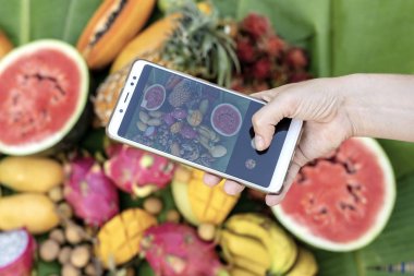 Kadın eli tropikal meyvelerin fotoğrafını çekiyor. Kahvaltıda çekilmiş akıllı telefon fotoğrafı. Tatlı mango, papaya, pitahaya, muz, karpuz, ananas. Çiğ vejetaryen sağlıklı yiyecekler
