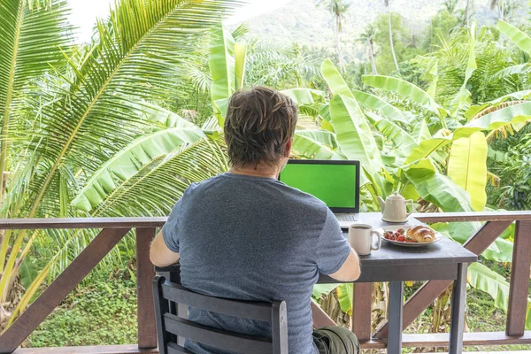 Old man working at laptop at terrace in tropical home during breakfast. Business concept.